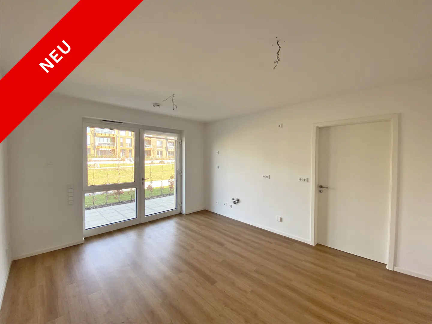 Bright, empty room with wood floor, white walls, and a glass door to the outside. Red "NEU" banner in the corner.