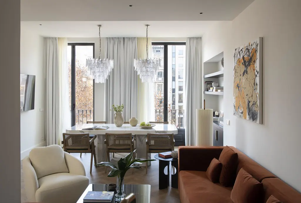Bright, modern living and dining area with a rust-colored sofa, white armchair, and marble dining table under two crystal chandeliers.