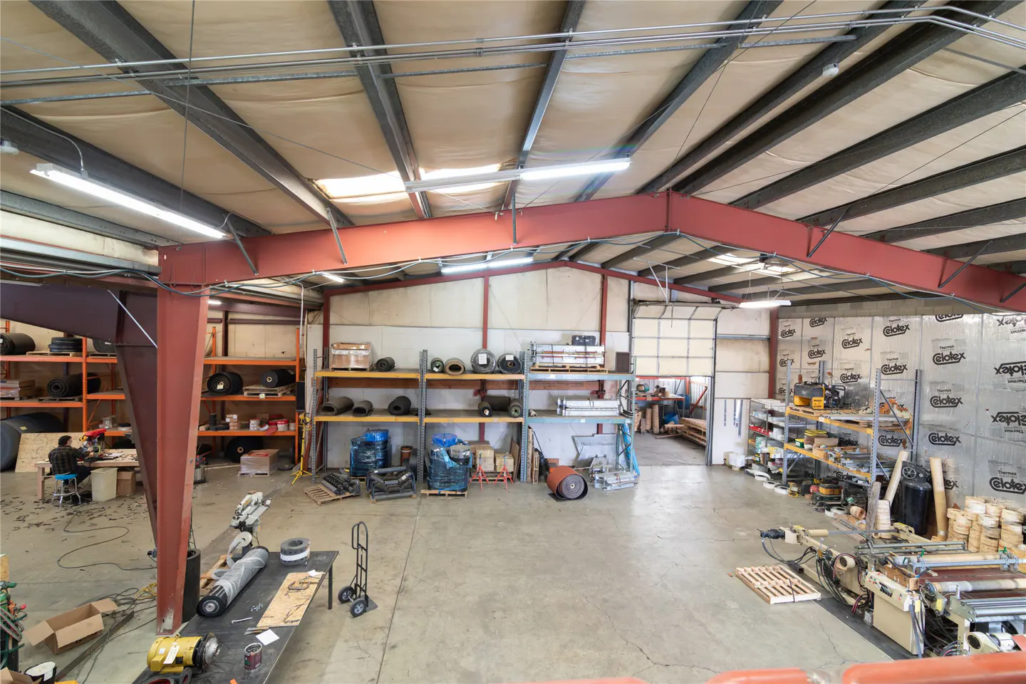 Wide shot of a warehouse interior with red steel beams, shelves, equipment, and a worker in the background.