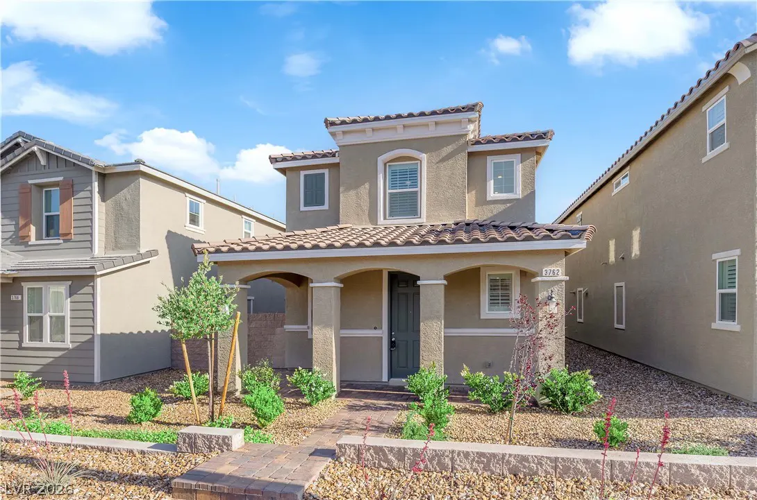Two-story beige house with a tiled roof, arched porch, and green door. Landscaped front yard with brick walkway and blue sky.