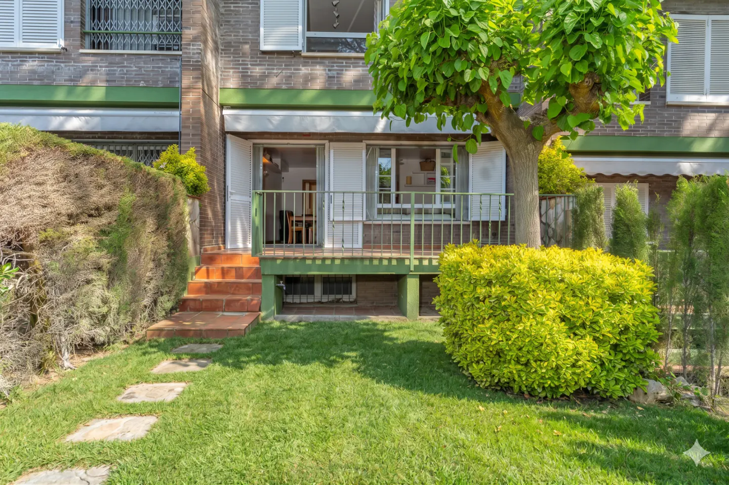 Exterior view of a brick townhouse with a green lawn, stone path, and a small porch with white railings and open shutters.