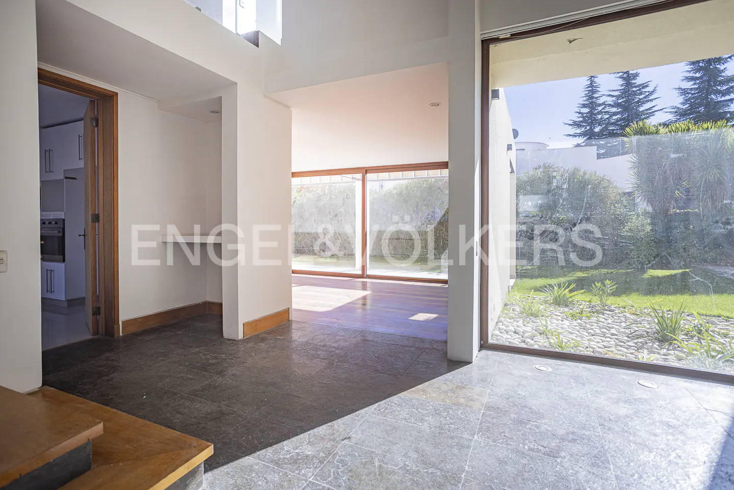 Interior view of a modern home with gray tile floors, white walls, and large windows overlooking a green lawn and trees. A doorway leads to a kitchen.