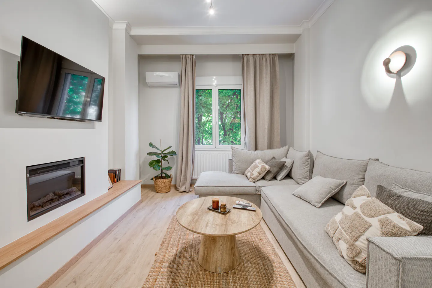 Living room with gray sectional sofa, round wood table on jute rug, TV over fireplace, and window with green trees outside.