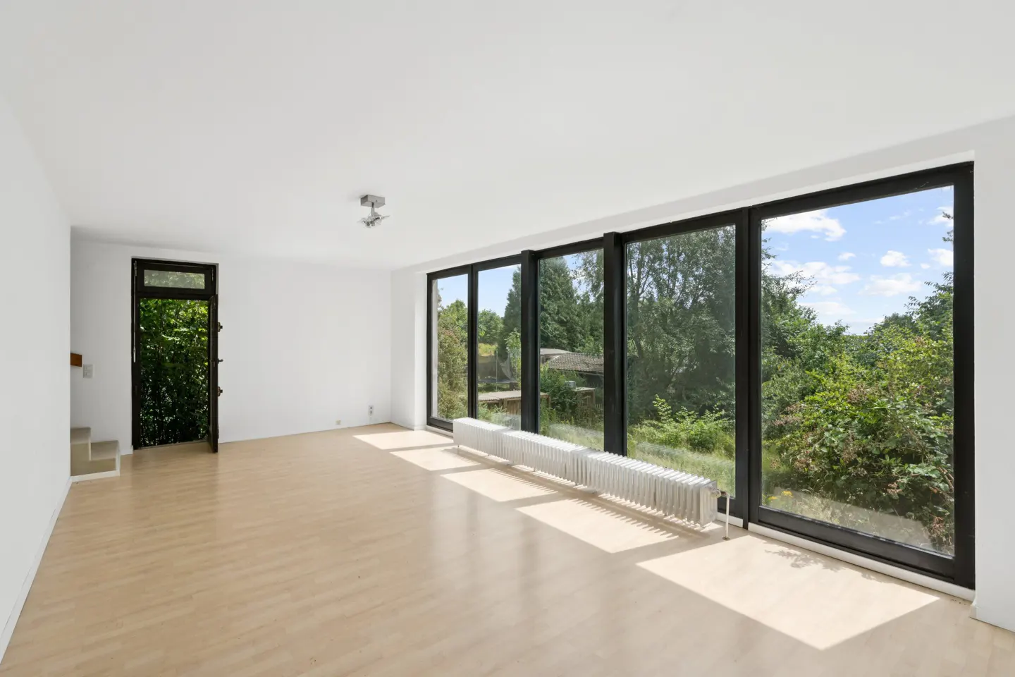 Bright, empty room with light wood floors, white walls, and black-framed windows overlooking lush greenery. A white radiator sits beneath the windows.