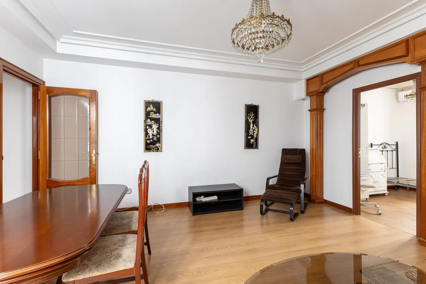 A bright living room with a wood dining table, brown chair, and a crystal chandelier. Two black and gold wall hangings decorate the white walls.