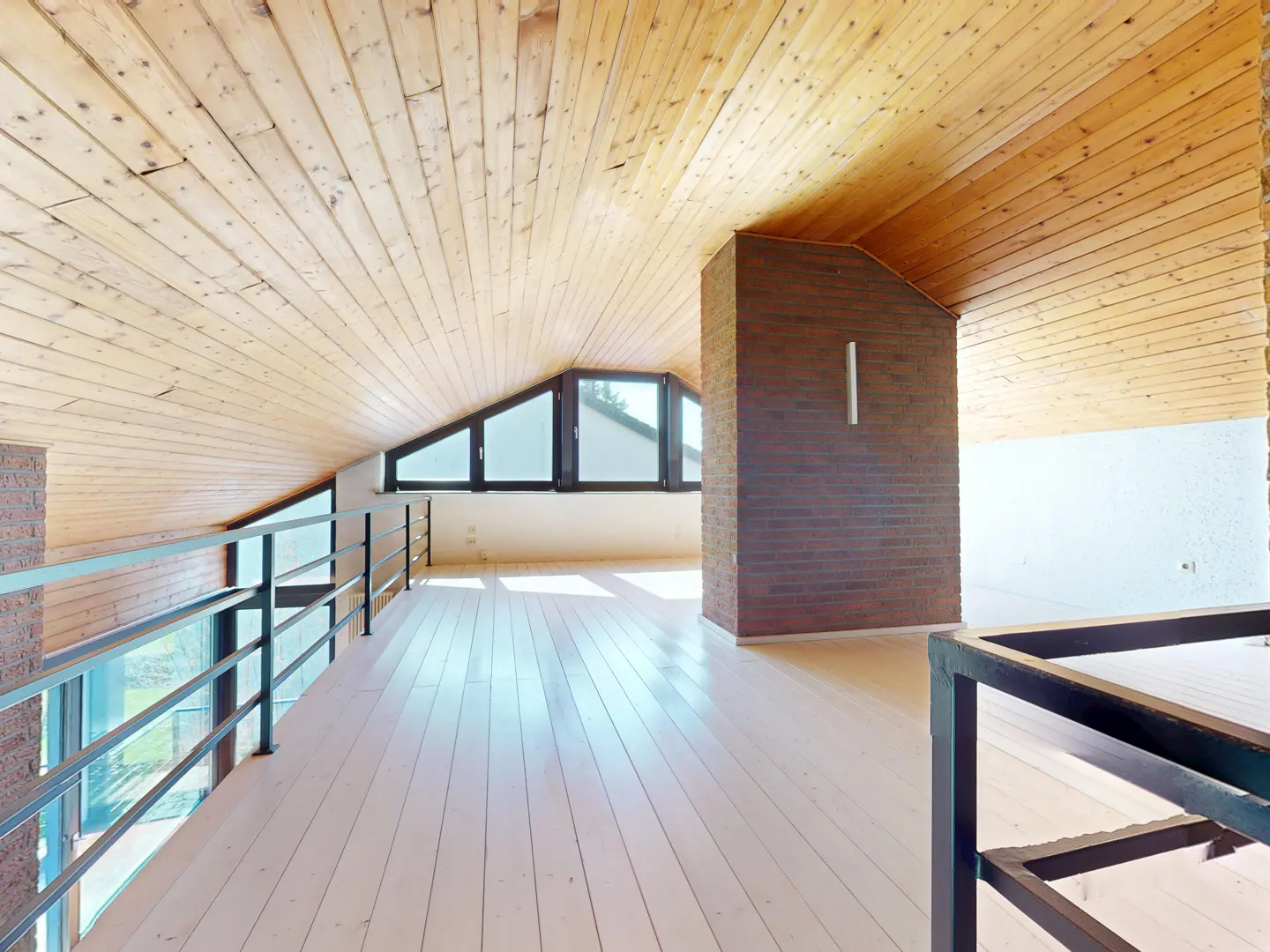 Attic room with light wood ceiling, white wood floor, and brick accent wall. Black metal railing and triangular windows.