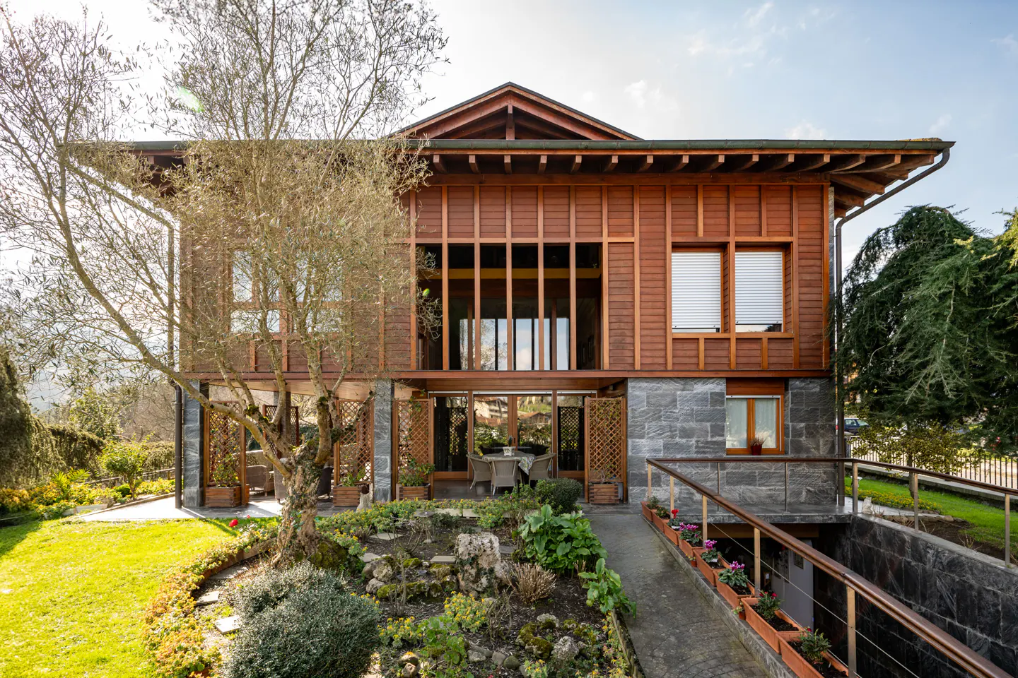 Two-story house with wood siding and stone base, surrounded by lush greenery and a garden. A walkway leads to a lower level entrance.