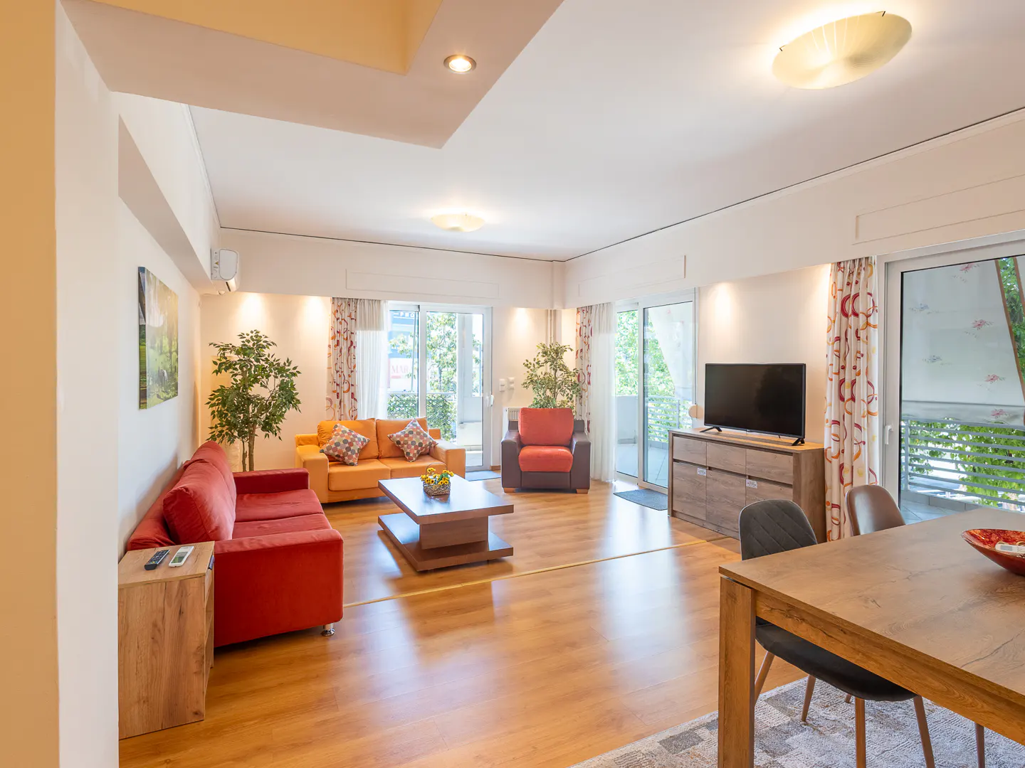 Bright living room with a red sofa, orange couch, armchair, wooden coffee table, TV, and dining area with wood flooring.
