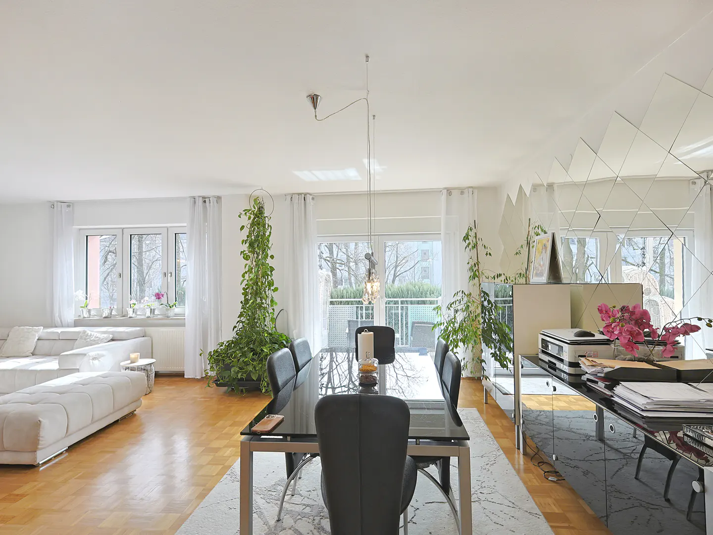 Bright, modern dining room with a black glass table, black chairs, and a white sofa. Large windows and plants add natural light and greenery.