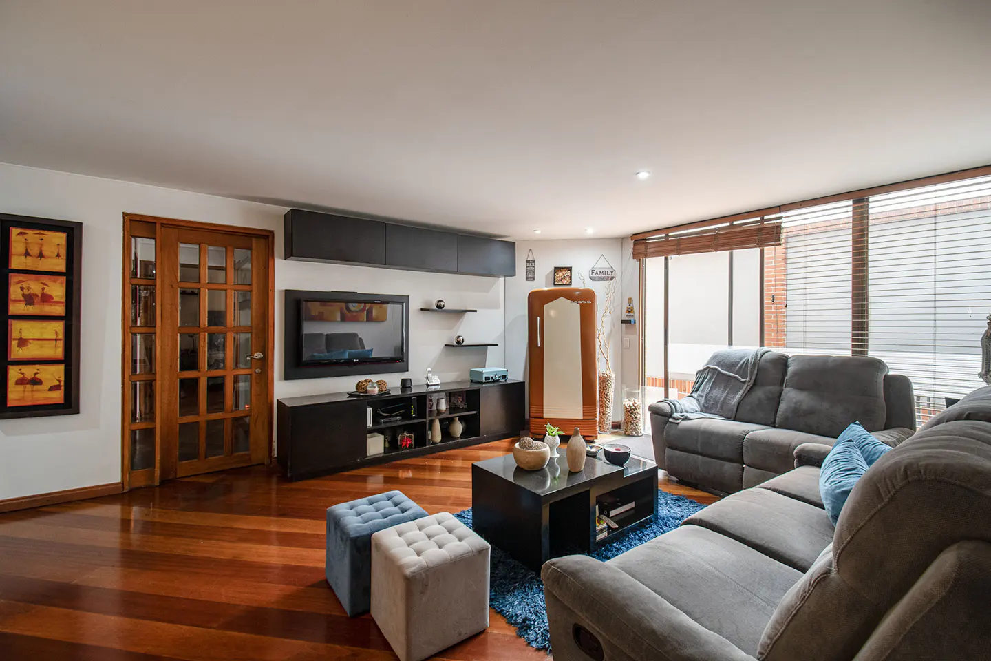 Living room with hardwood floors, gray sofas, and a black entertainment center. A blue rug and ottomans add color. Large windows provide natural light.