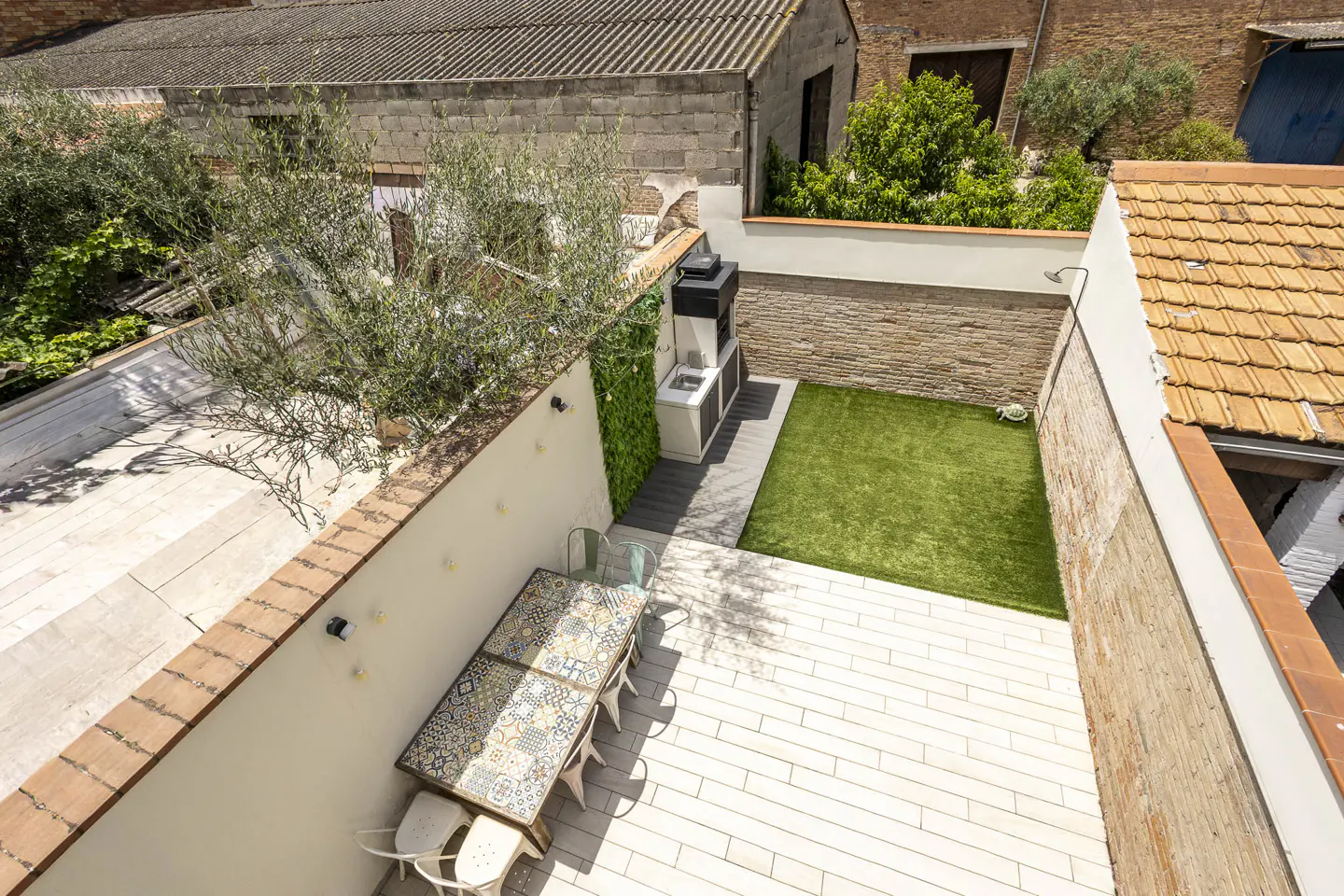High-angle view of a patio with a tiled dining table, outdoor grill, artificial grass area, and surrounding brick walls.
