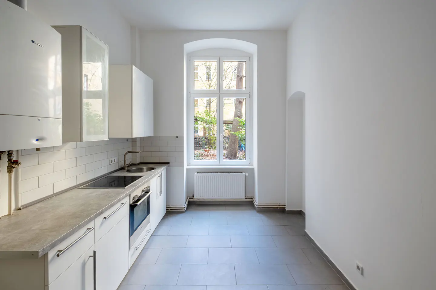 Bright, empty kitchen with white cabinets, gray counters and tile floor. A window looks out onto a courtyard with trees.