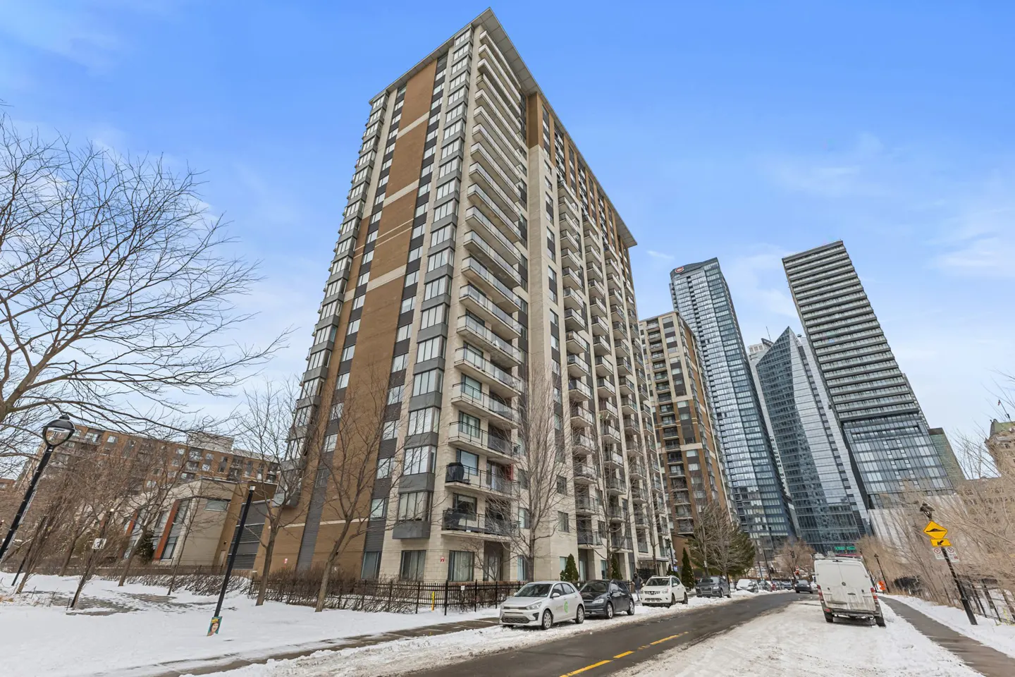 Exterior view of a tall brick apartment building with balconies, set against a backdrop of modern skyscrapers and a snowy street.