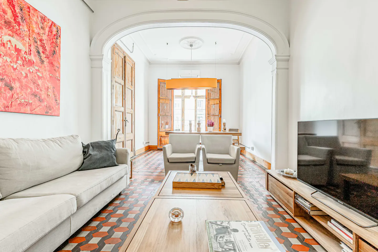 Bright living room with a beige sofa, wooden coffee table, and a TV. An archway leads to a dining area with wooden shutters. Red abstract art on the wall.