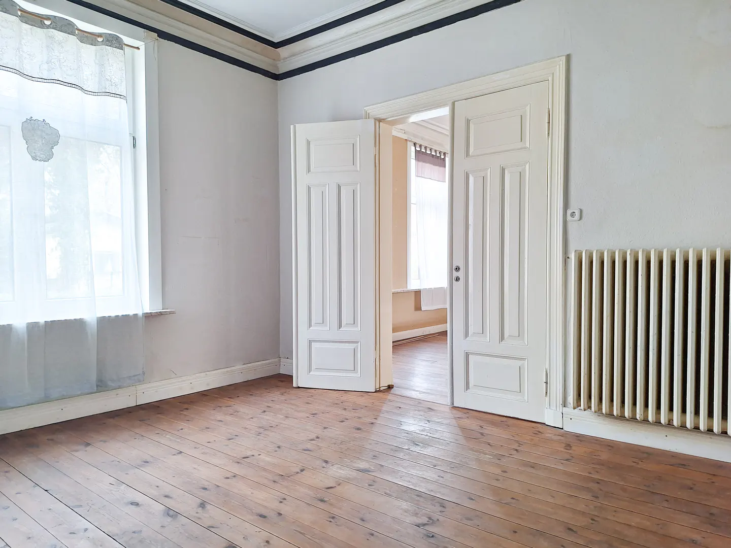 Empty room with wood floors, white walls, and an open white double door. A window with sheer curtains is on the left, and a radiator is on the right.