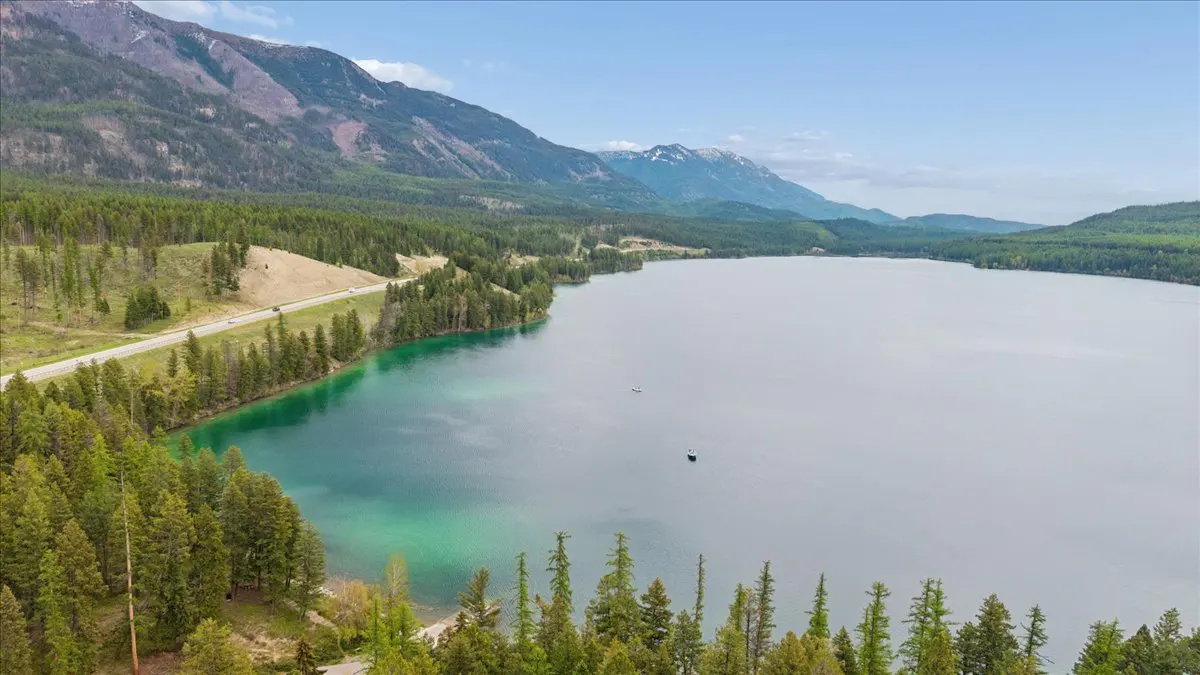 Aerial view of a turquoise lake bordered by a dense pine forest, a winding road, and majestic mountains under a blue sky.