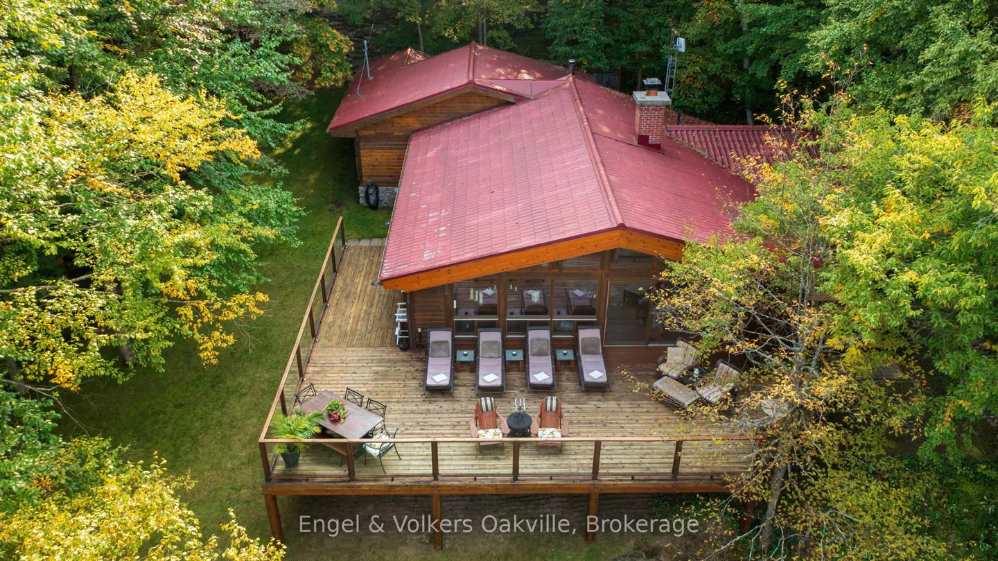 Aerial view of a log cabin with a red roof, surrounded by trees. A large wooden deck features lounge chairs and a dining table.