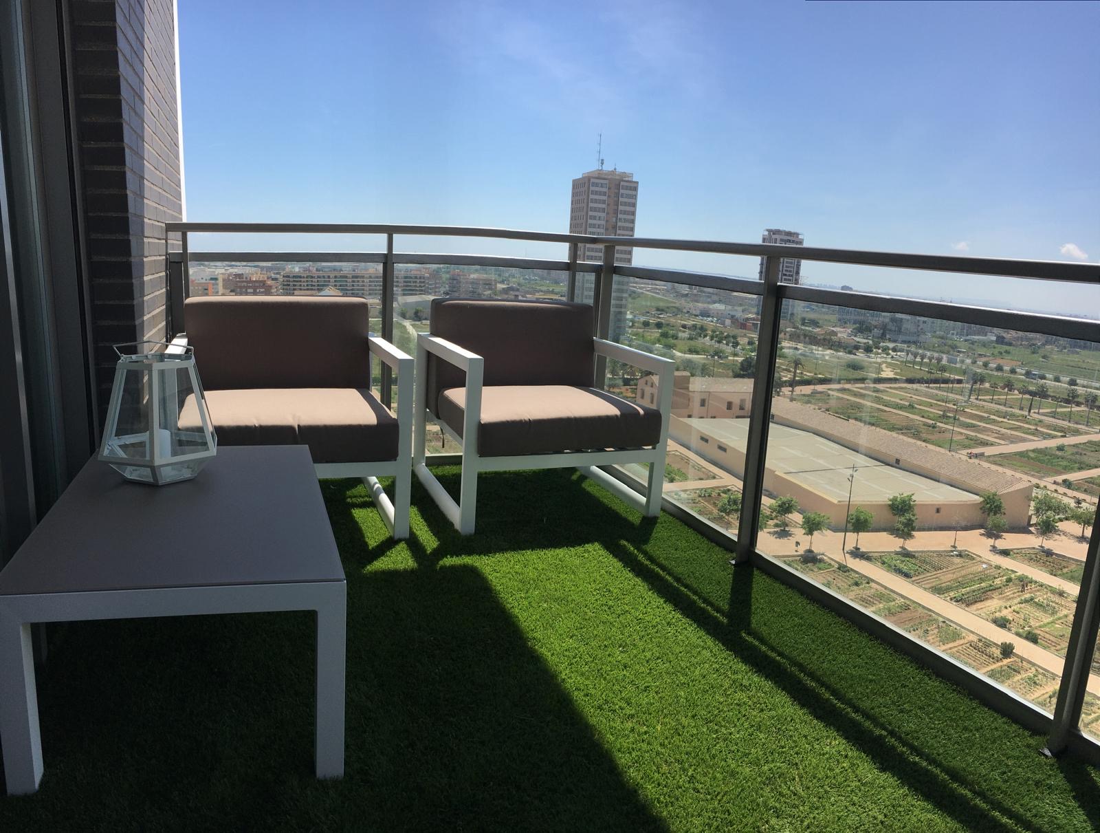 Modern balcony with two brown chairs, a small table, and artificial grass, overlooking a city landscape under a blue sky. [AI-generated text]
