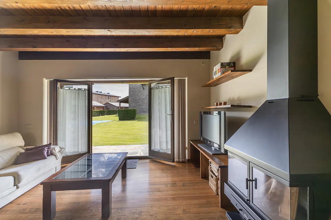 Living room with wood beams, white sofa, glass coffee table, modern fireplace, and open doors to a sunny green garden.