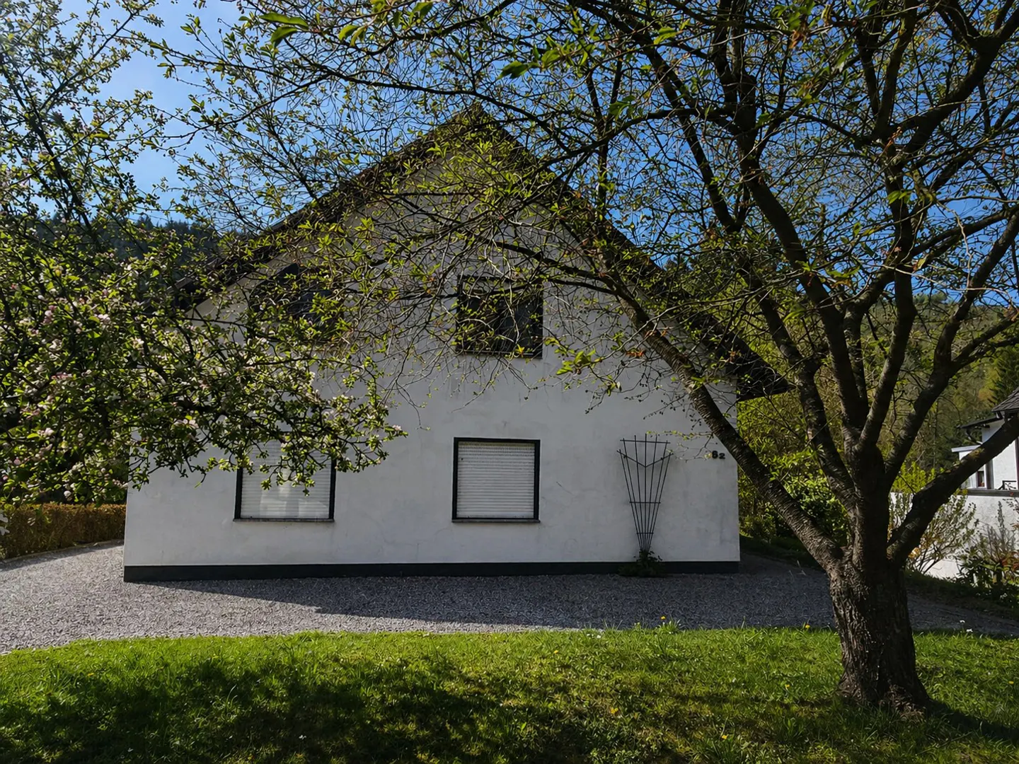 A white, two-story house with a black roof is framed by trees with green leaves. A green lawn is in front of the house.