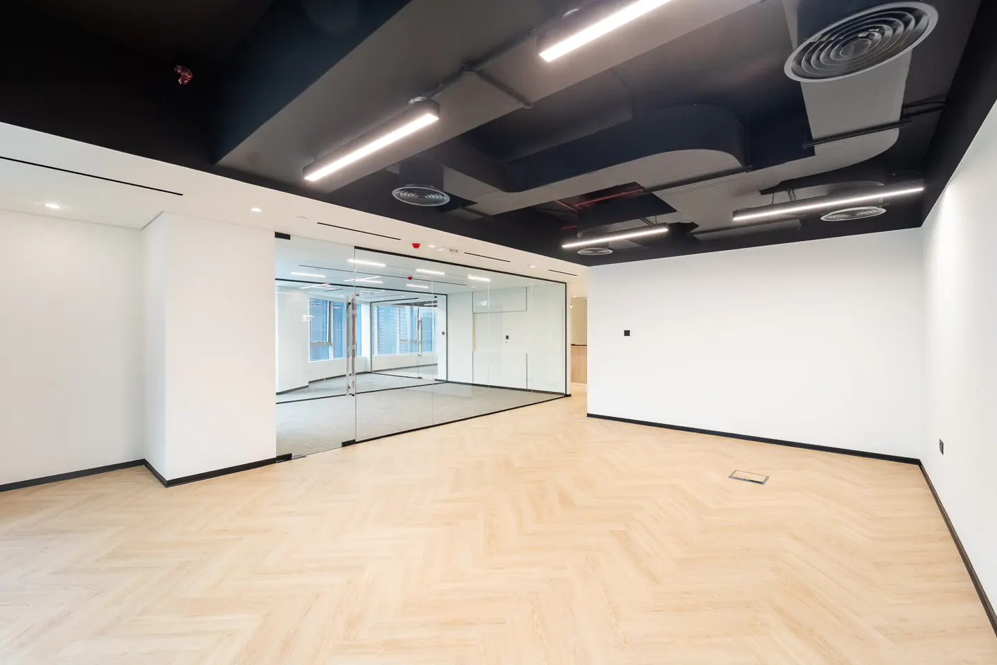 Empty office space with herringbone wood floors, white walls, and a black ceiling with linear lights and vents. A glass-walled room is visible.