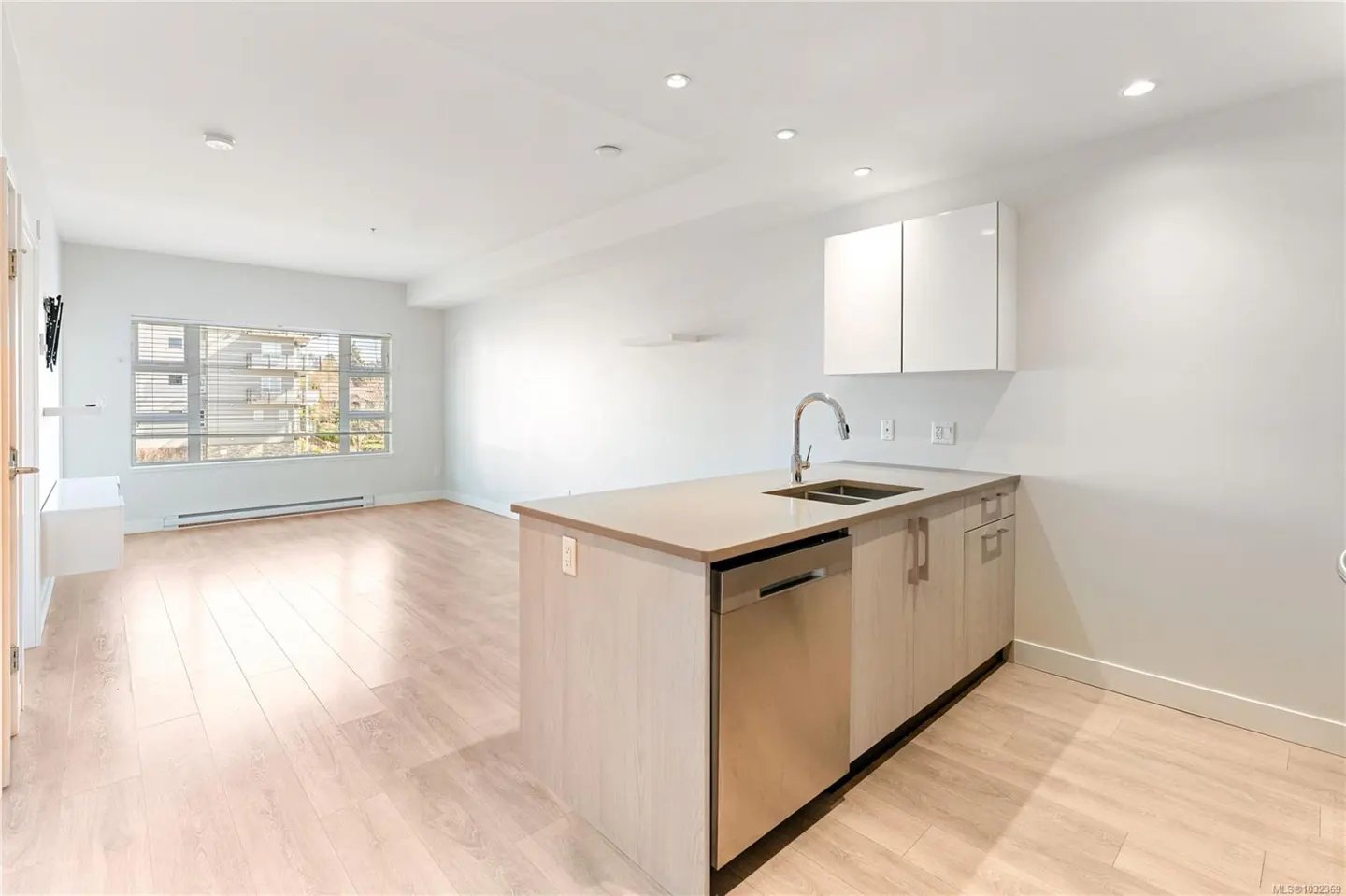 Bright, open-concept apartment with light wood floors, white walls, and a kitchen island with stainless steel appliances. A large window overlooks a residential area.