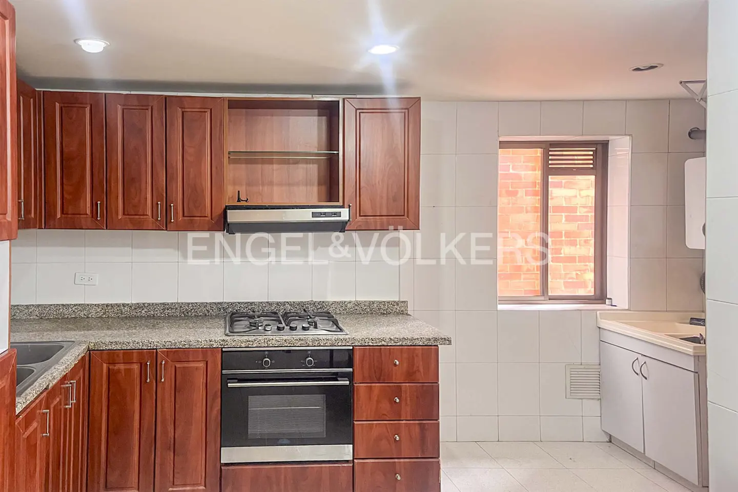 A kitchen with brown cabinets, granite countertops, and white tiled walls. A window shows a brick building outside.