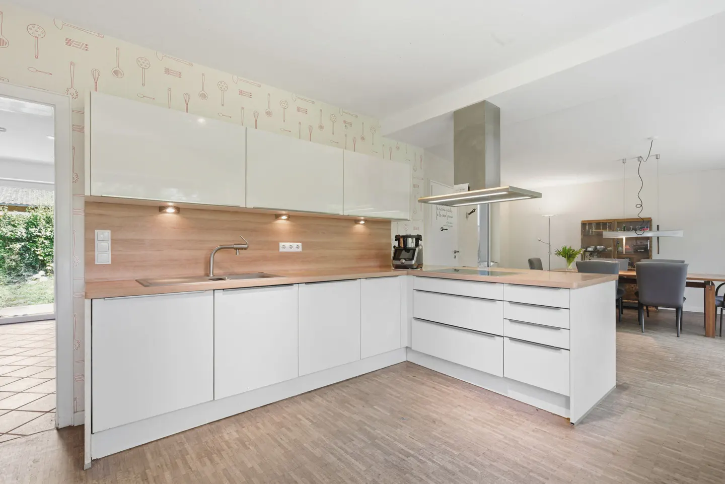 Bright, modern kitchen with white cabinets, wood countertops, and stainless steel appliances. Dining area visible in the background.