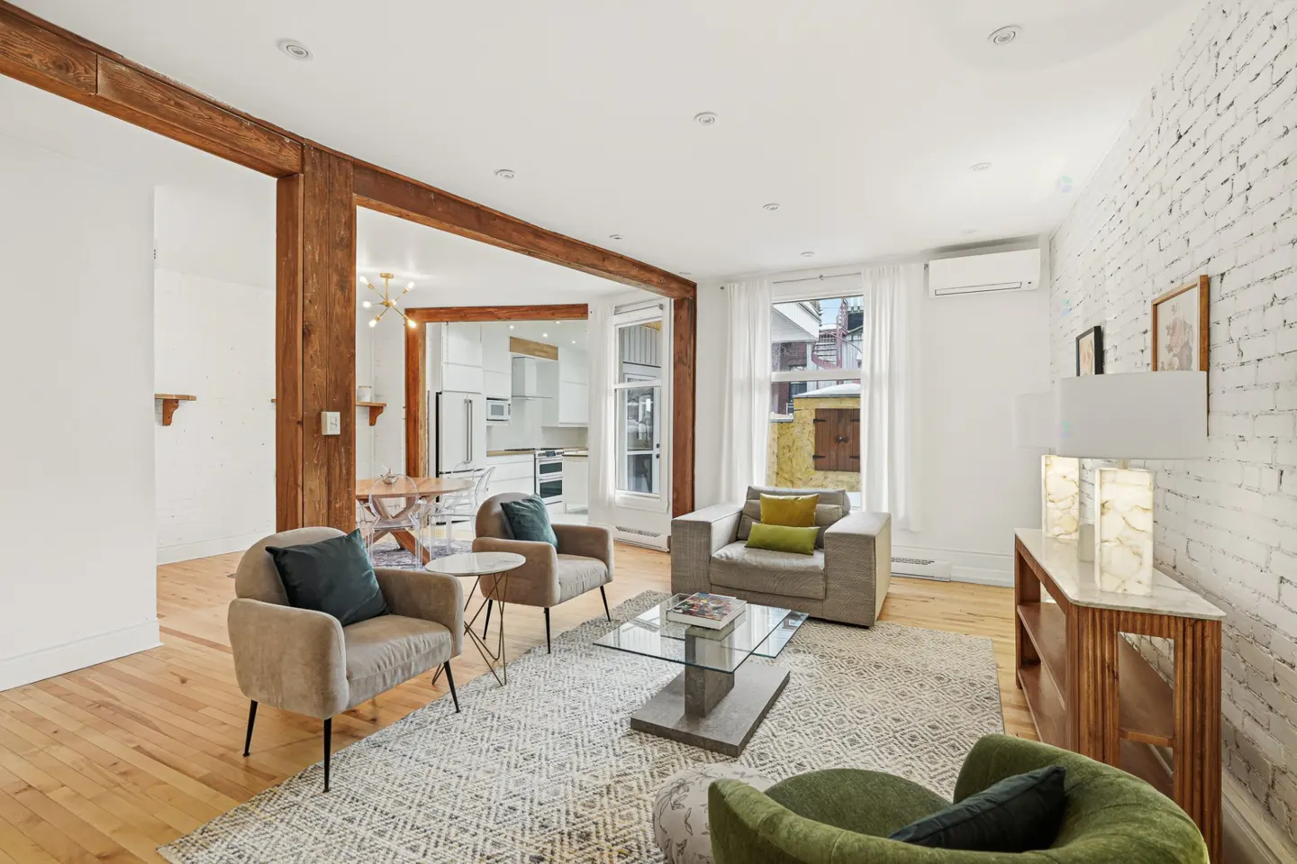 Bright living room with hardwood floors, exposed wood beams, and a white brick wall. Several chairs and a glass coffee table sit on a patterned rug.