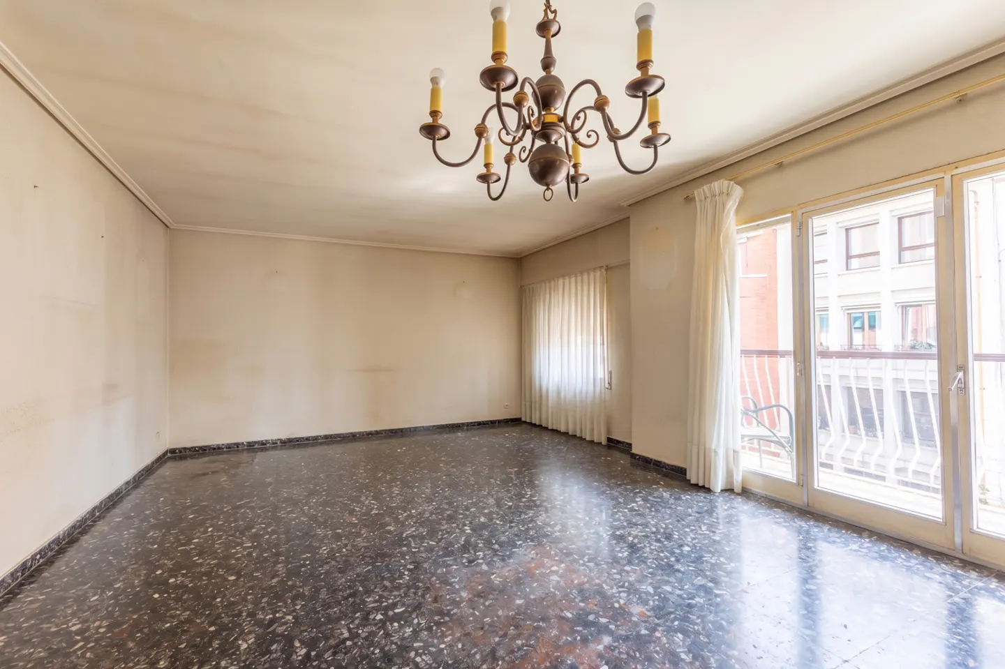 Empty room with dark speckled floor, beige walls, and a bronze chandelier. Balcony doors with white curtains lead to a city view.