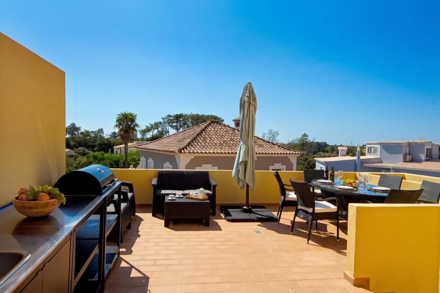 Outdoor patio with a grill, dining table set for a meal, and lounge seating under a bright blue sky.