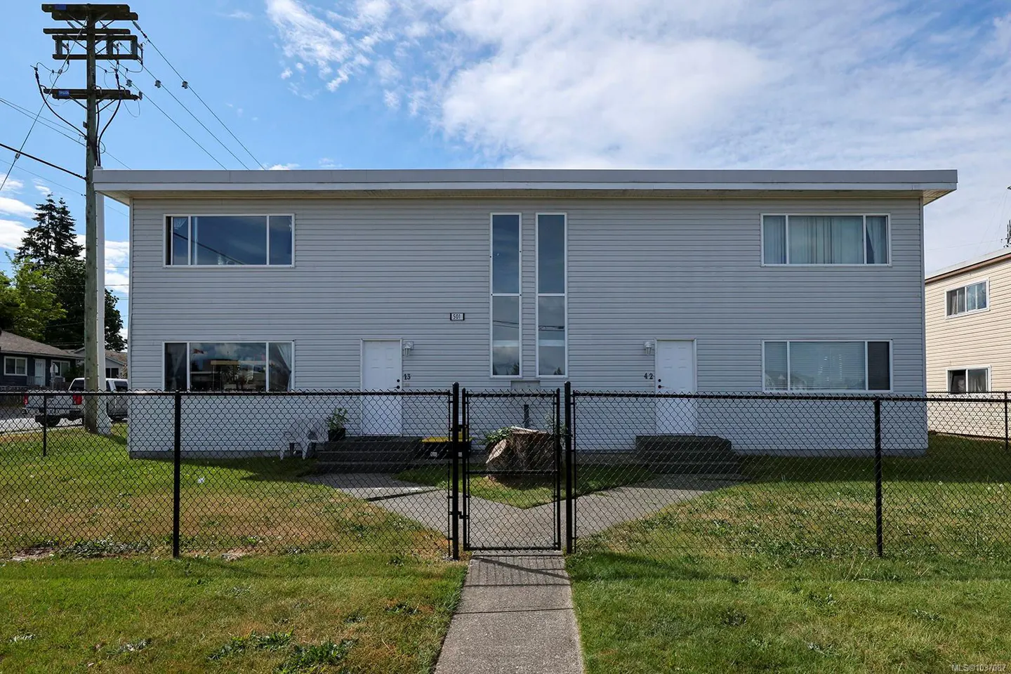 Two-story, light gray duplex with a black chain-link fence, green lawn, and a sidewalk leading to a gate.