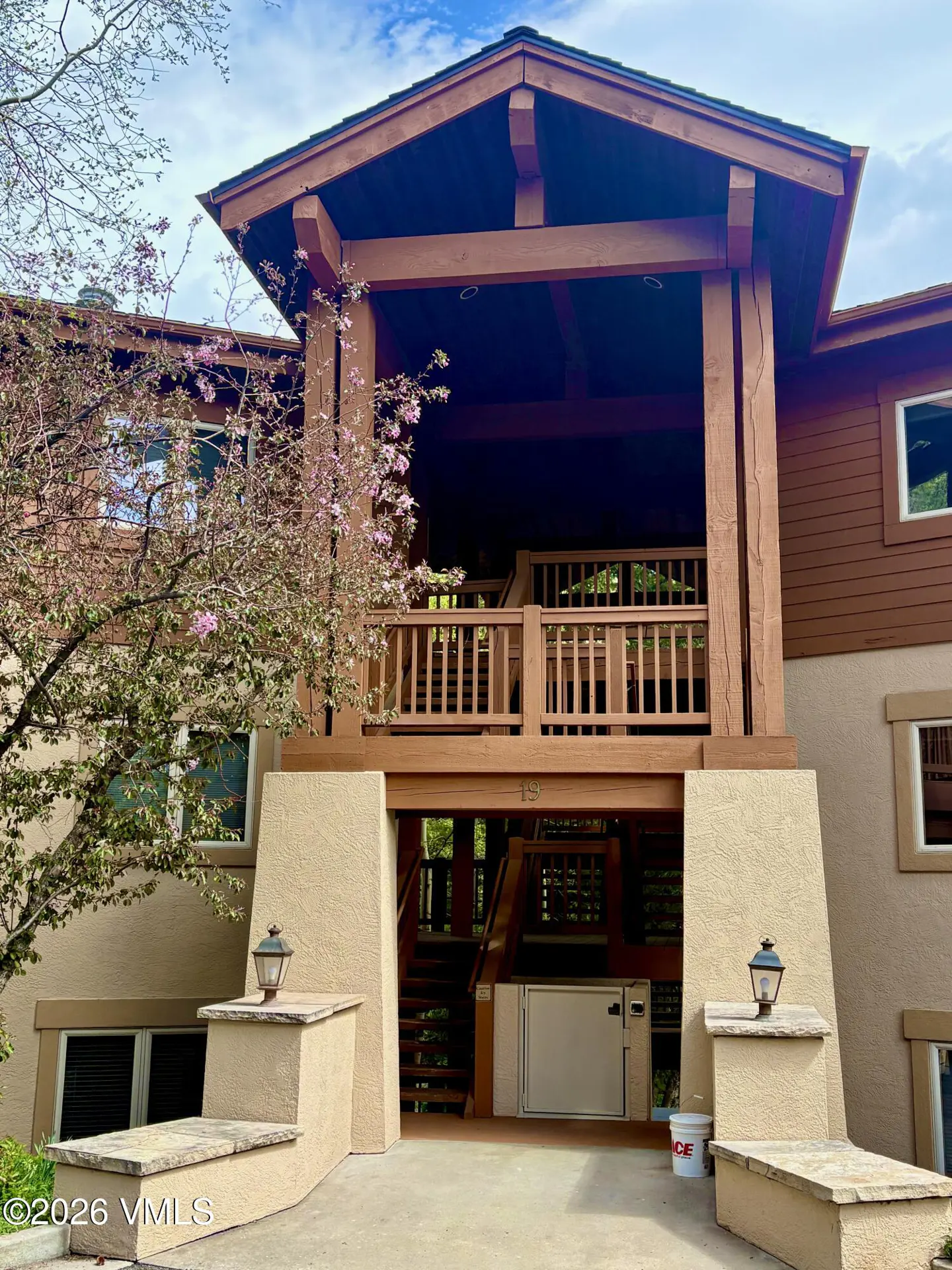 Two-story condo with brown wood trim, beige stucco, and a covered balcony. A flowering tree is on the left.