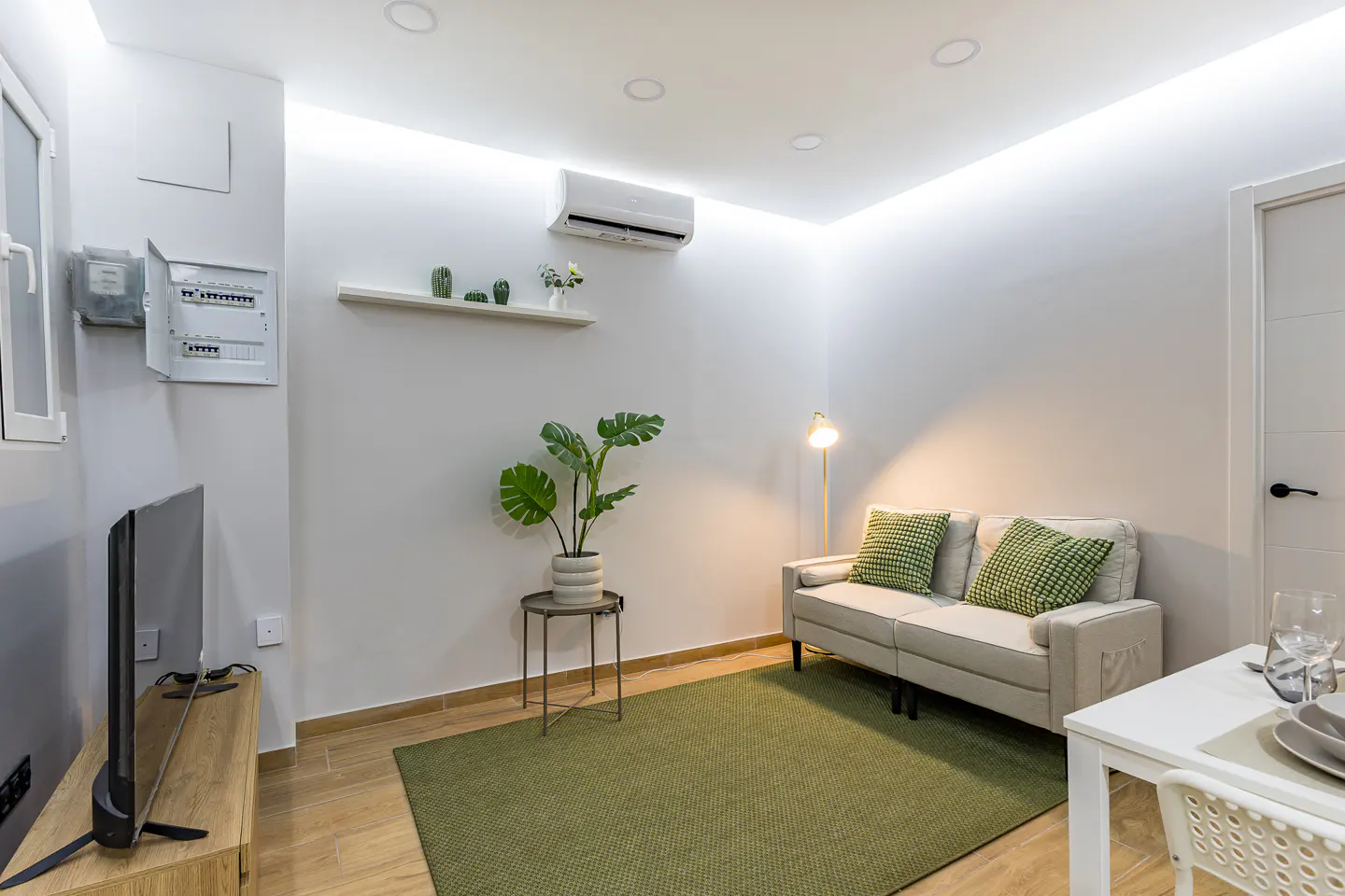 Bright living room with a beige sofa, green rug, and a potted plant on a side table. A white dining table is on the right.