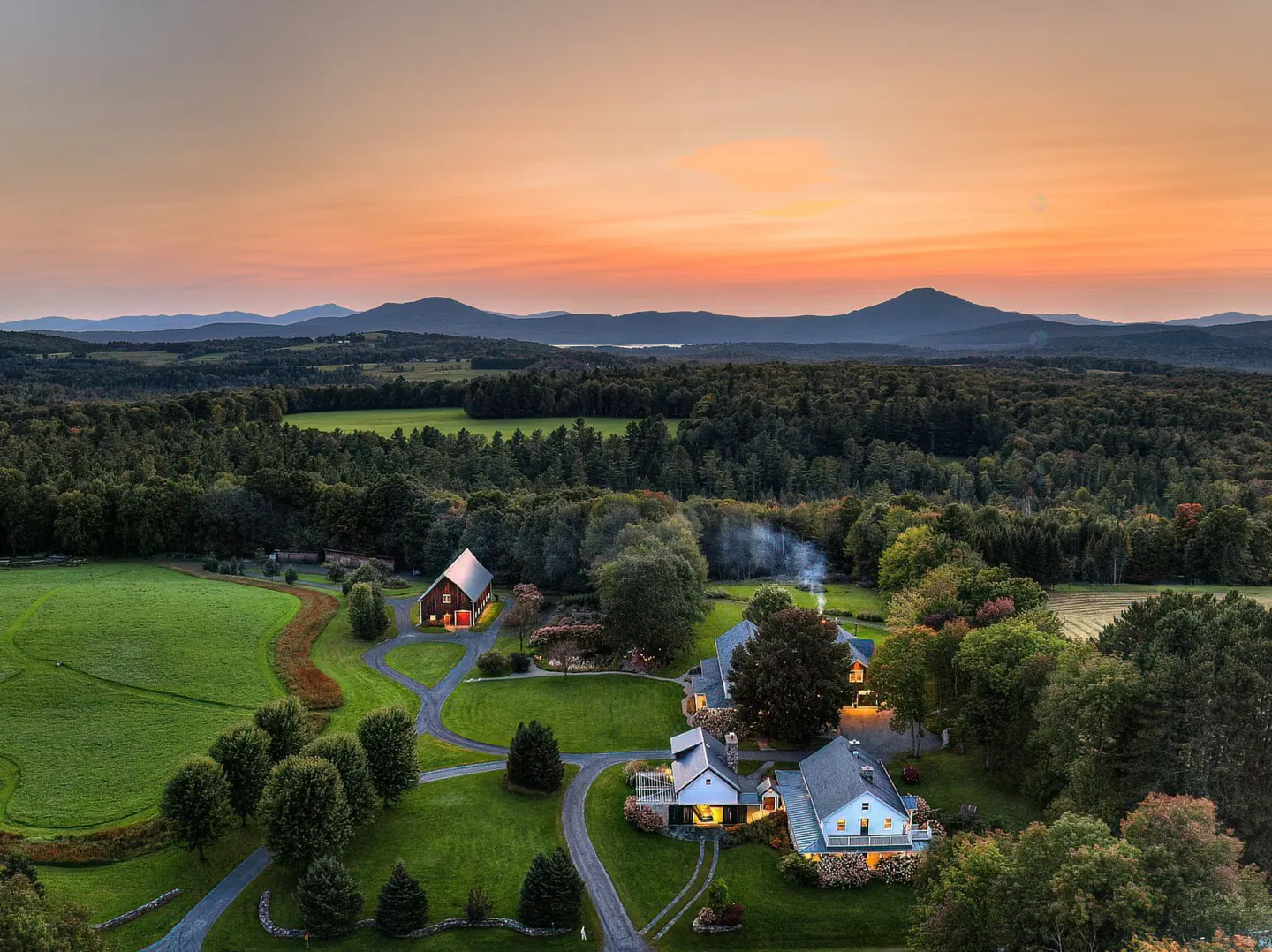 Aerial view of a rural estate with a red barn, white houses, green lawns, and a forest backdrop under a colorful sunset sky.