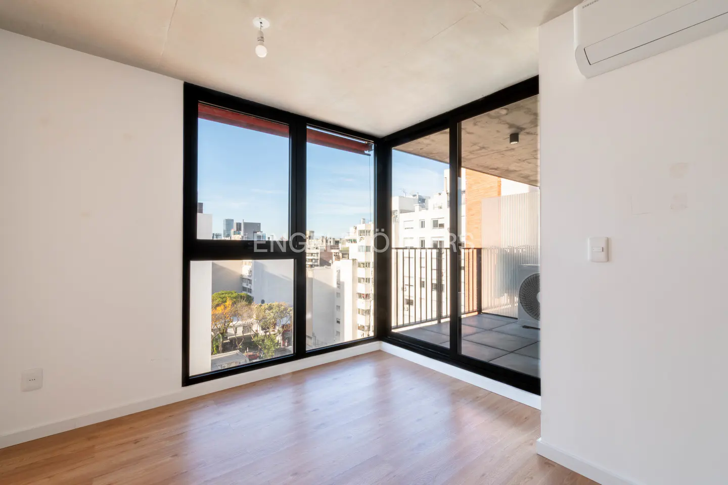 Bright, empty room with wood floors, white walls, and a large black-framed window overlooking a city skyline and balcony.