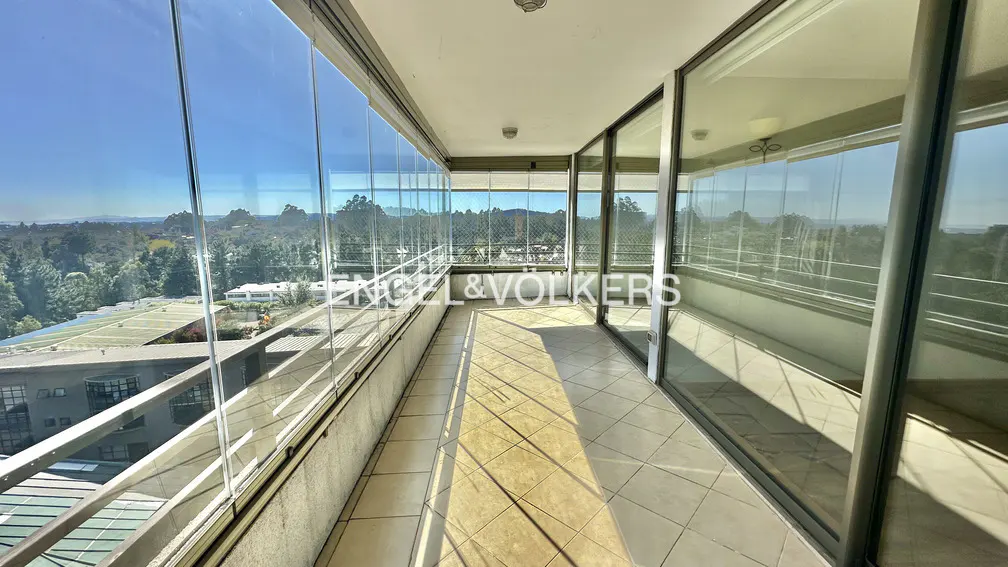A sunlit balcony with glass walls offers a panoramic view of trees and a blue sky. The floor is tiled in a light color.