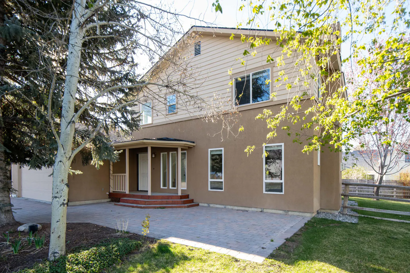 Two-story house with tan siding and brown stucco, a covered porch, and a stone patio. Trees frame the house.