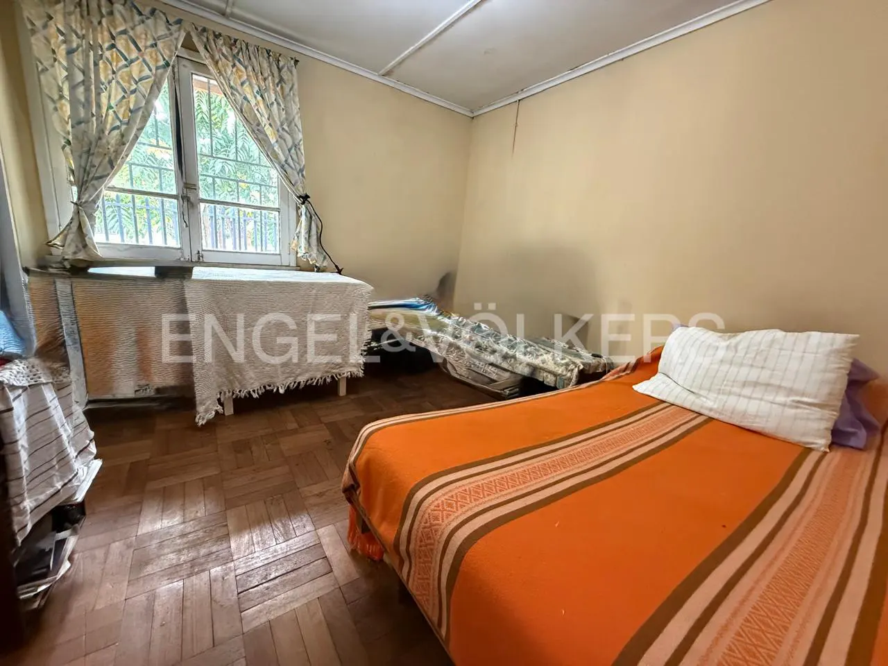 Bedroom with hardwood floors, beige walls, and two beds. One bed has an orange striped blanket. Window with patterned curtains.