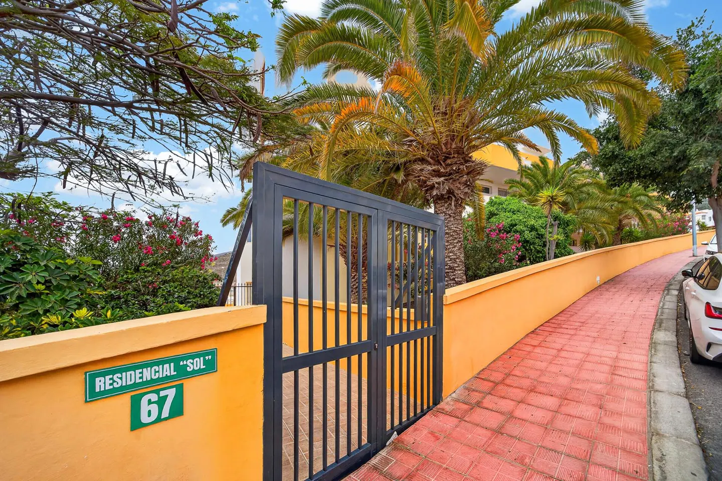 Entrance to Residencial "Sol" 67, with a black metal gate and orange wall. Palm trees and a red brick path add to the tropical feel.
