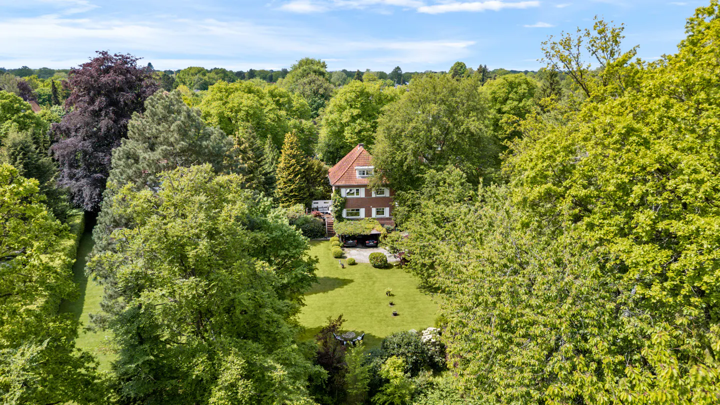 Aerial view of a three-story house with a red tile roof, surrounded by lush green trees and a blue sky.