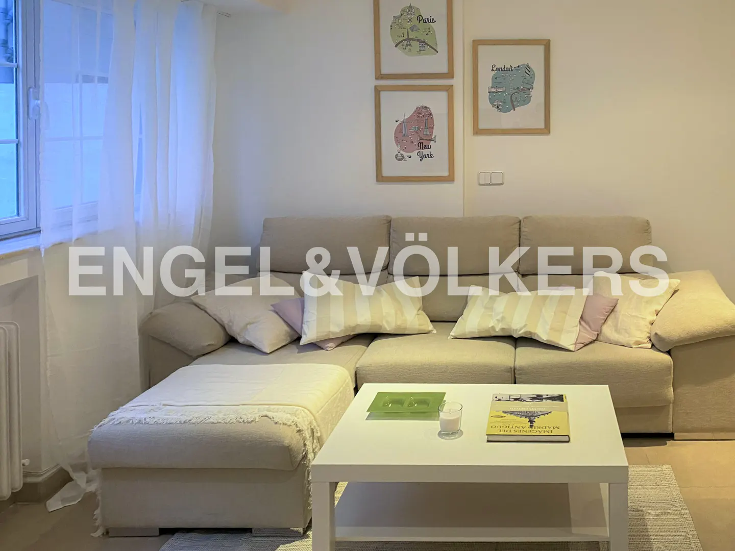 Living room with beige sectional sofa, white coffee table, and framed city map art on the wall. Natural light filters through sheer curtains.