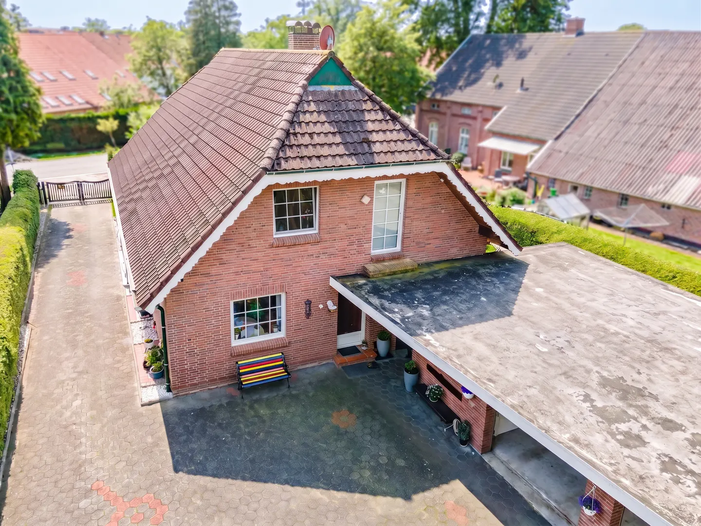 Aerial view of a two-story brick house with a red tile roof and a carport. A colorful bench sits near the front door.
