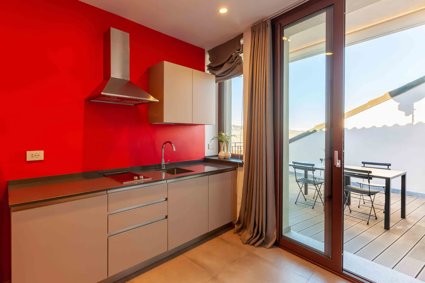 Modern kitchen with red wall, beige cabinets, stainless steel hood, and sliding glass door to a balcony with table and chairs.