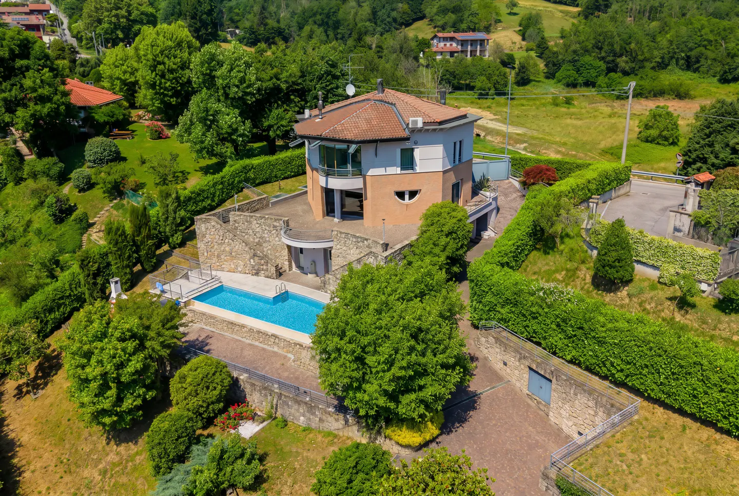 Aerial view of a two-story peach and white house with a swimming pool, surrounded by lush green trees and stone walls.