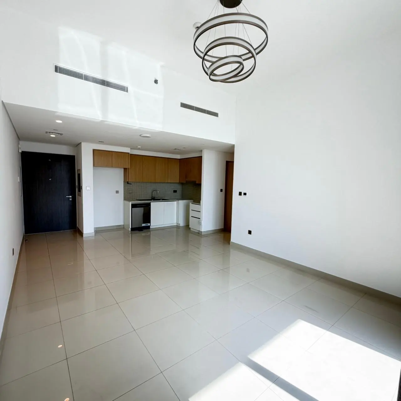 Bright, empty apartment with white walls, tile floors, and a modern kitchen. A circular chandelier hangs from the ceiling.