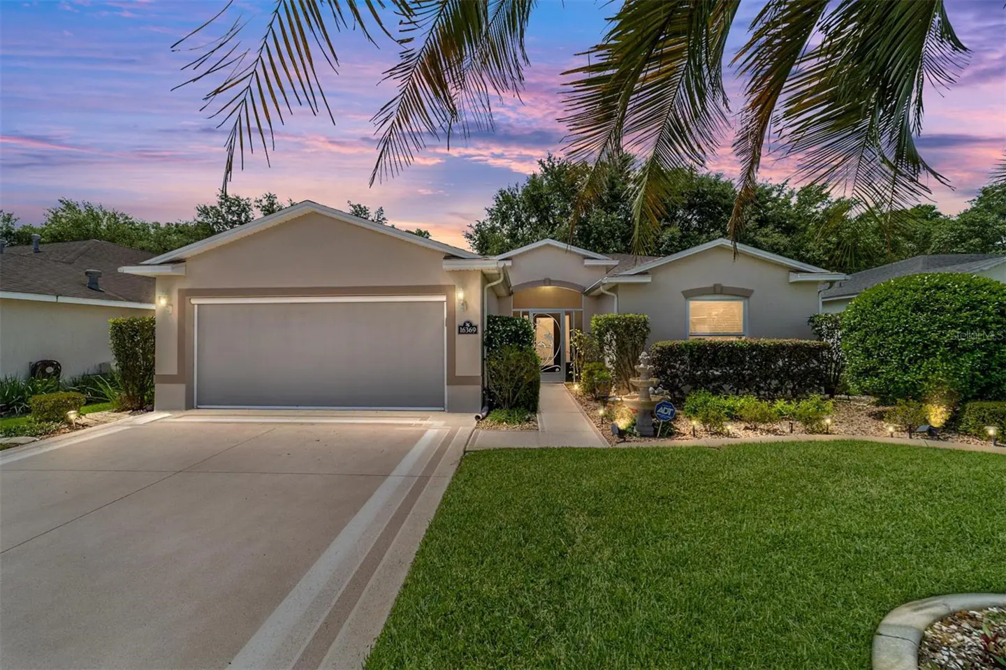 Beige single-story house with a gray garage door, green lawn, and palm trees against a colorful sunset sky.