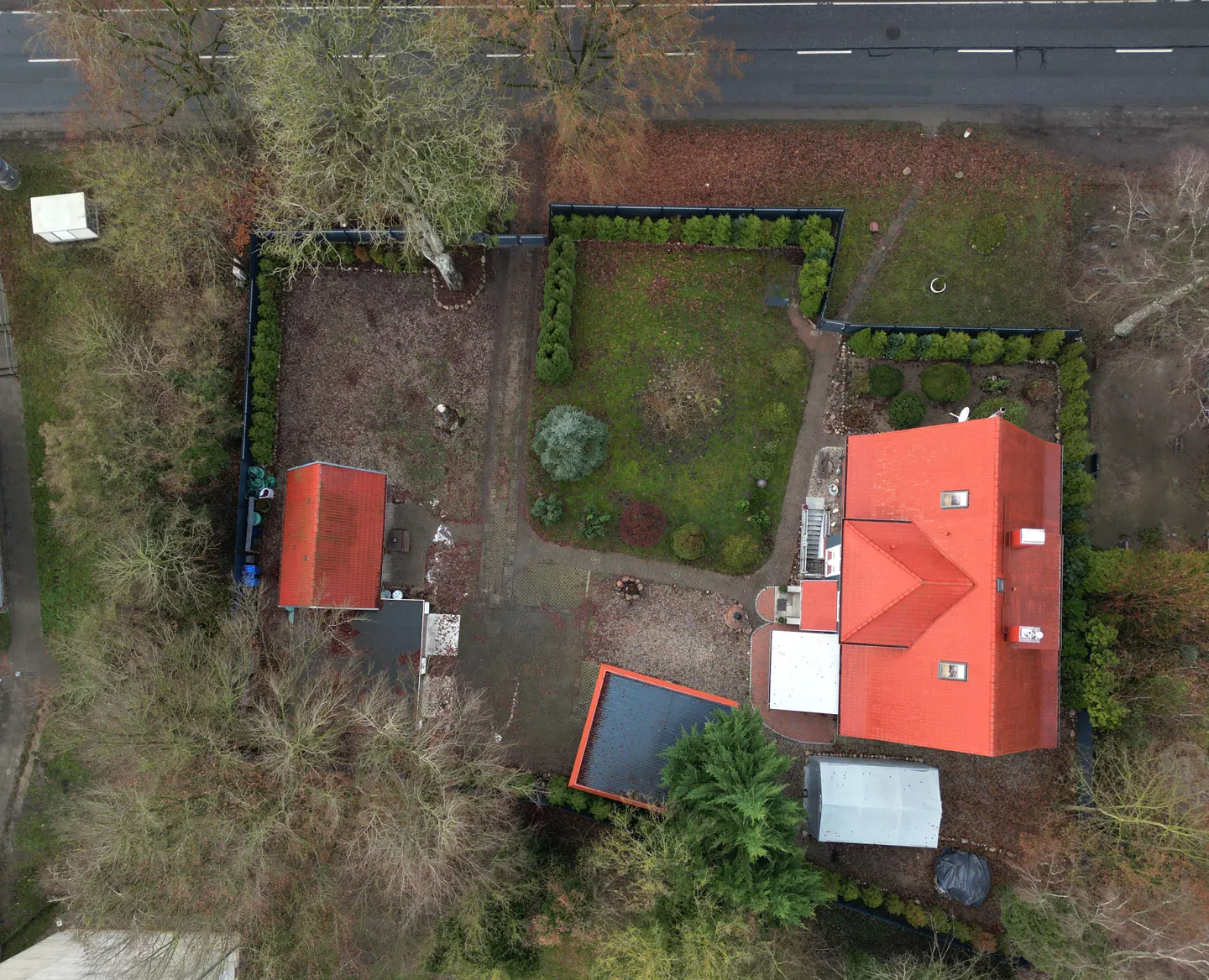 Aerial view of a red-roofed house with a green lawn, surrounded by trees and a black fence.