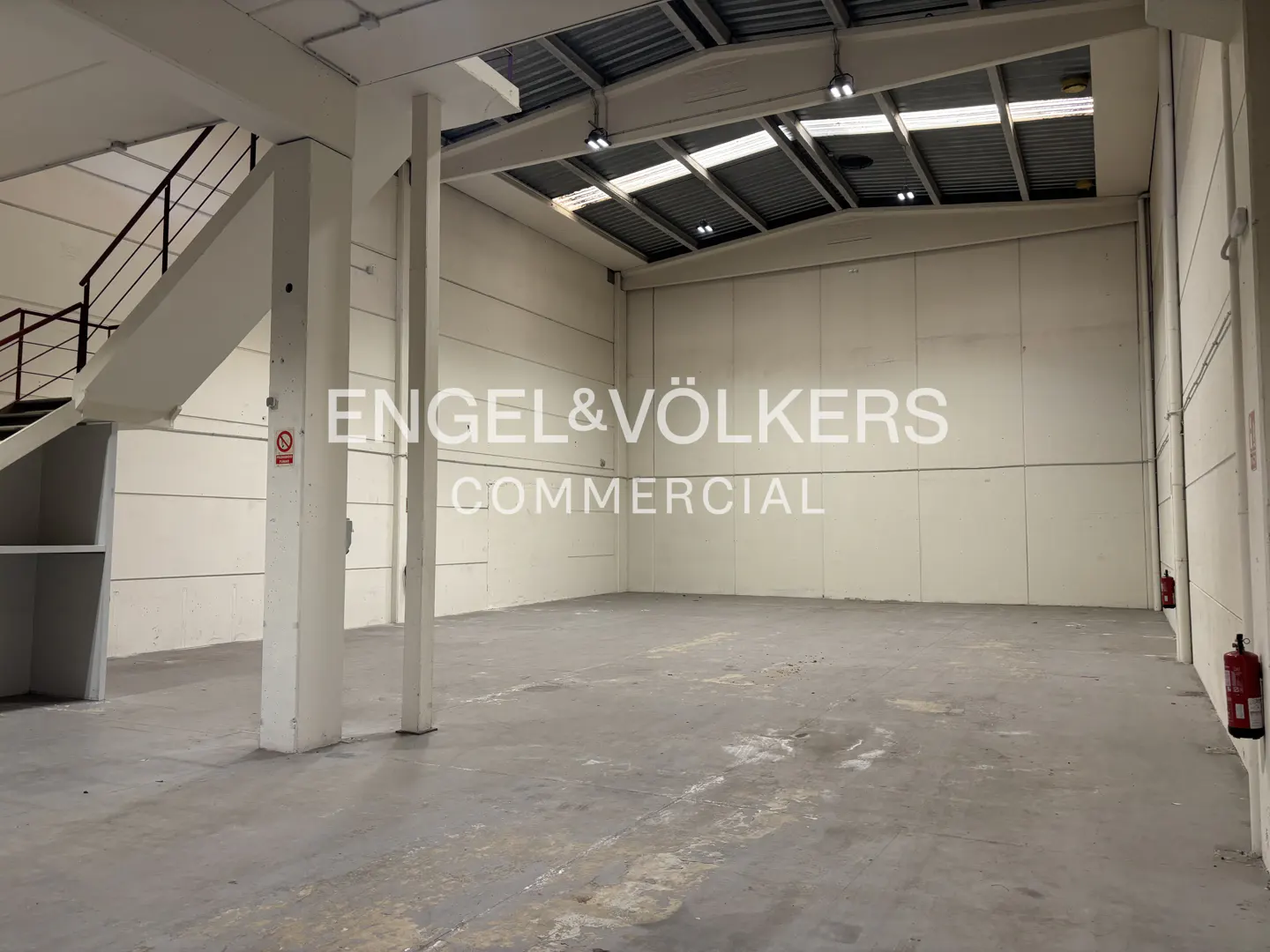 Empty commercial space with concrete floors, white walls, and metal ceiling. Stairs on the left and fire extinguishers on the right.