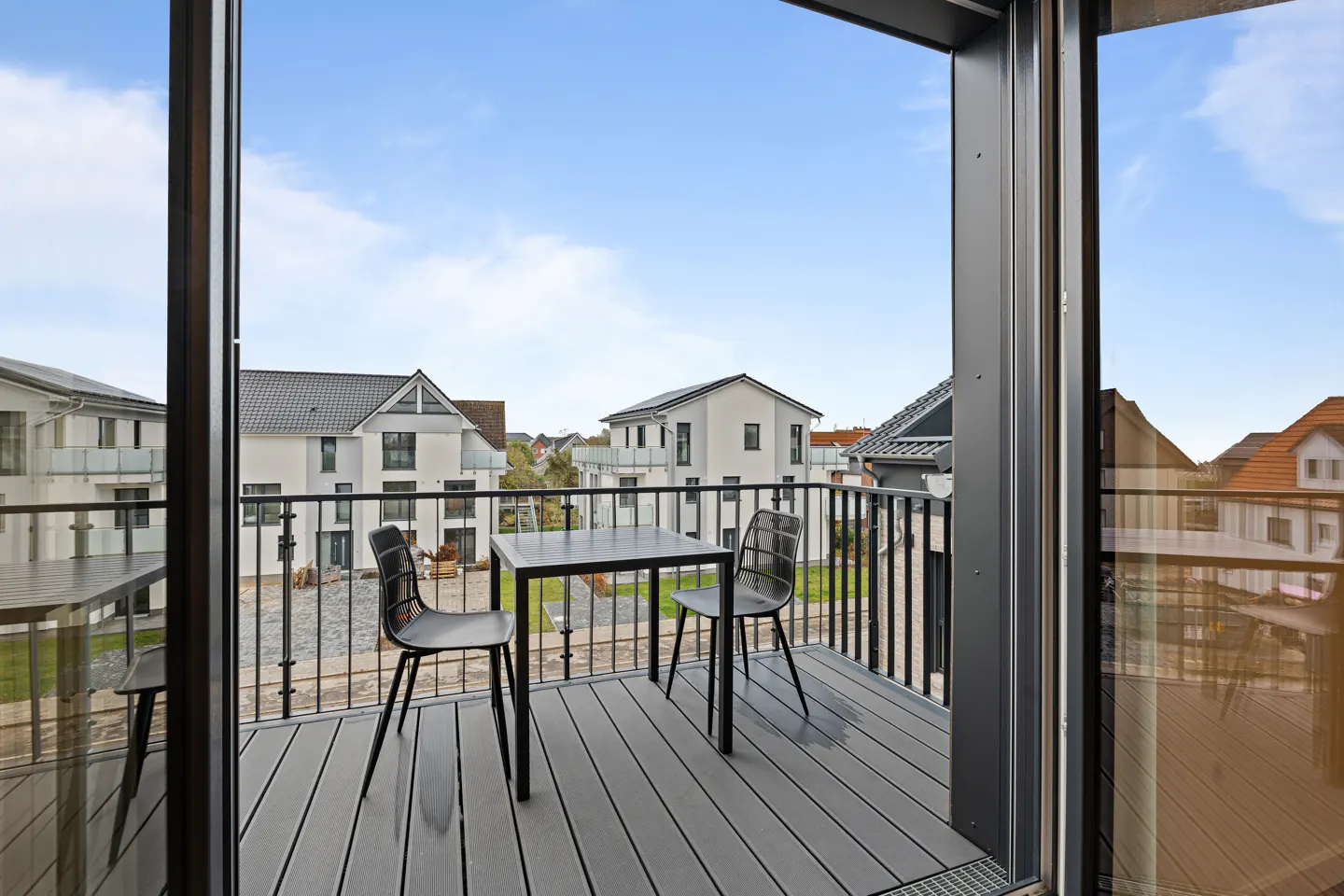 Balcony view with black table and chairs on gray wood deck, black railing, and houses in the background under a blue sky.